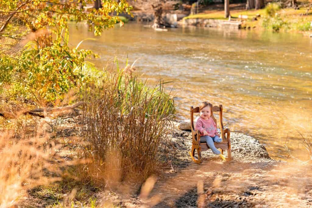 Beautiful fall scenery featuring a joyful child sitting on a wooden rocking chair by a river, surrounded by colorful autumn foliage.