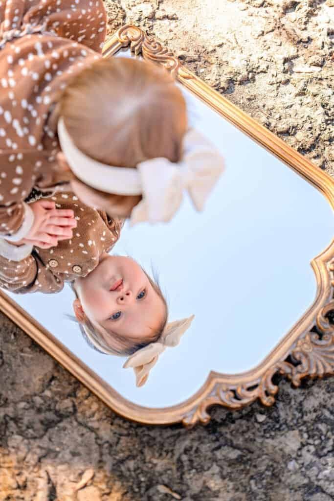 Child girl looking at her reflection in ornate vintage mirror outdoors.