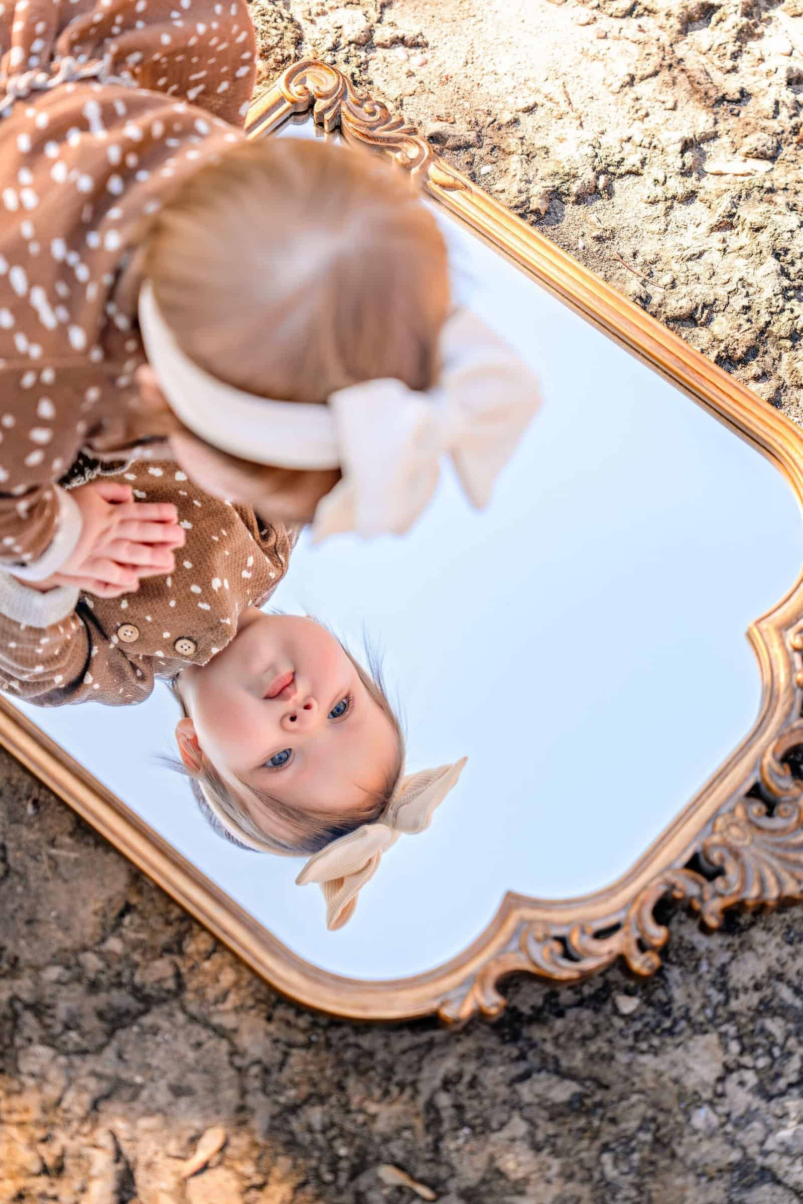 Child girl looking at her reflection in ornate vintage mirror outdoors.