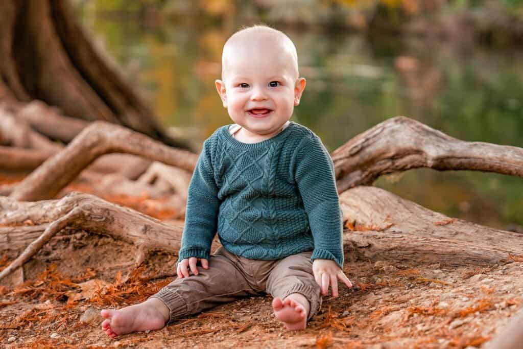 Baby sitting on fallen tree at park in autumn, smiling and looking at the camera, in a natural outdoor setting.