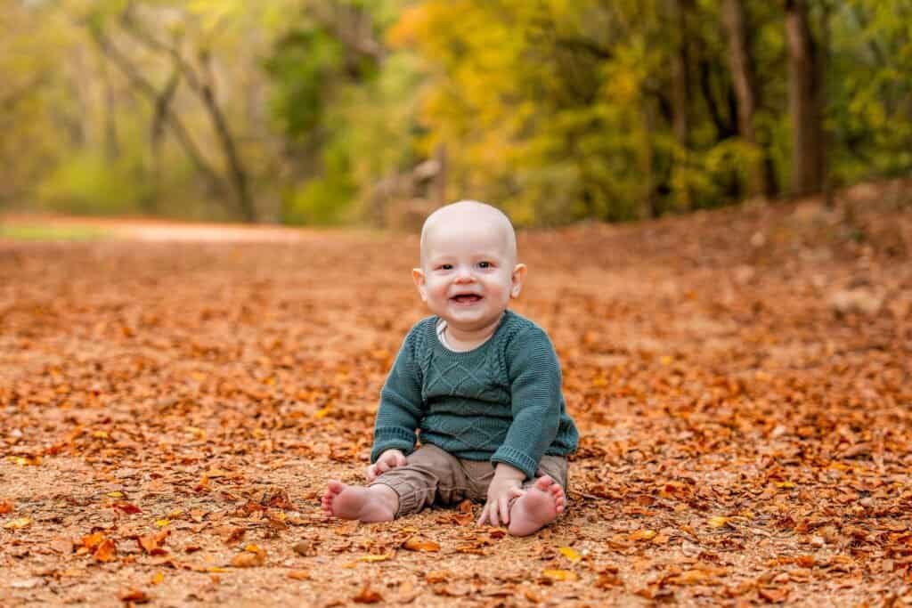Cute baby sitting on autumn fallen leaves in a park, colorful fall foliage background.