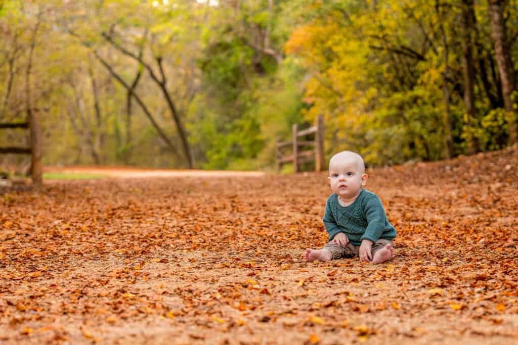 Adorable baby sitting outdoors on fallen leaves during fall, surrounded by colorful autumn trees.